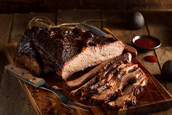 A sliced smoked brisket with a fork and sauce in the side and placed on a wooden board