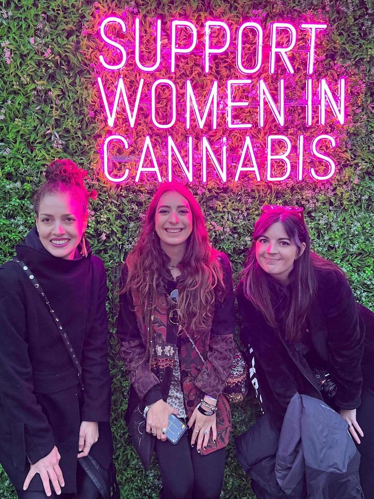Foto colorida de três mulheres na frente de um neon rosa dizendo:  “Apoie as mulheres na cannabis” 
