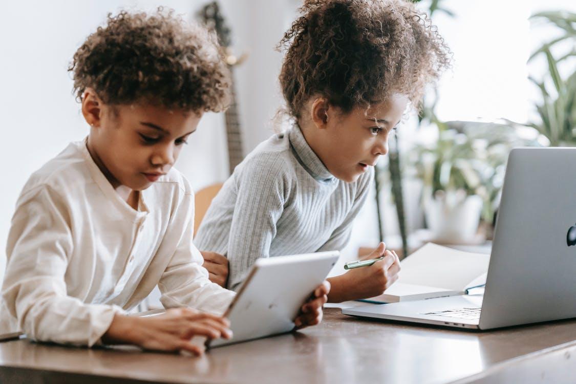 Focused African American children in casual clothes sitting at table and browsing laptop and tablet at home in daytime
