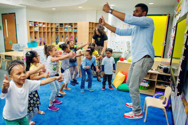 Adult male standing in front of multiple children with their arms stretched and thumbs up dancing on a blue carpet adult female standing on the side
