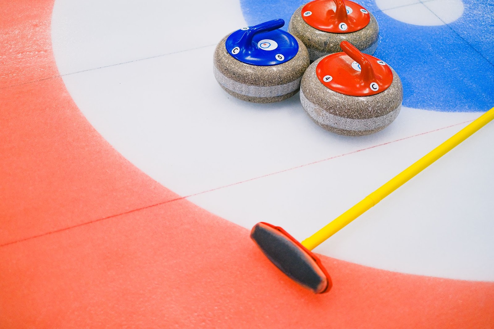 a curling broom with a yellow handle lying on the ice next to three rocks, two with red handles and one with a blue handle.