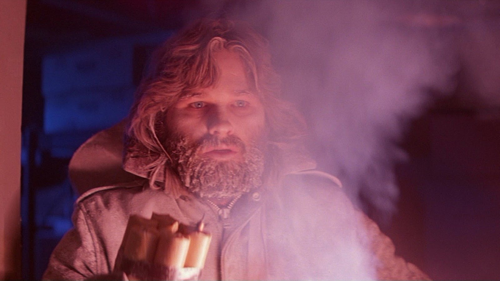A still of MacReady, played by Kurt Russell, with ice in his beard. There is smoke billowing in front of him.