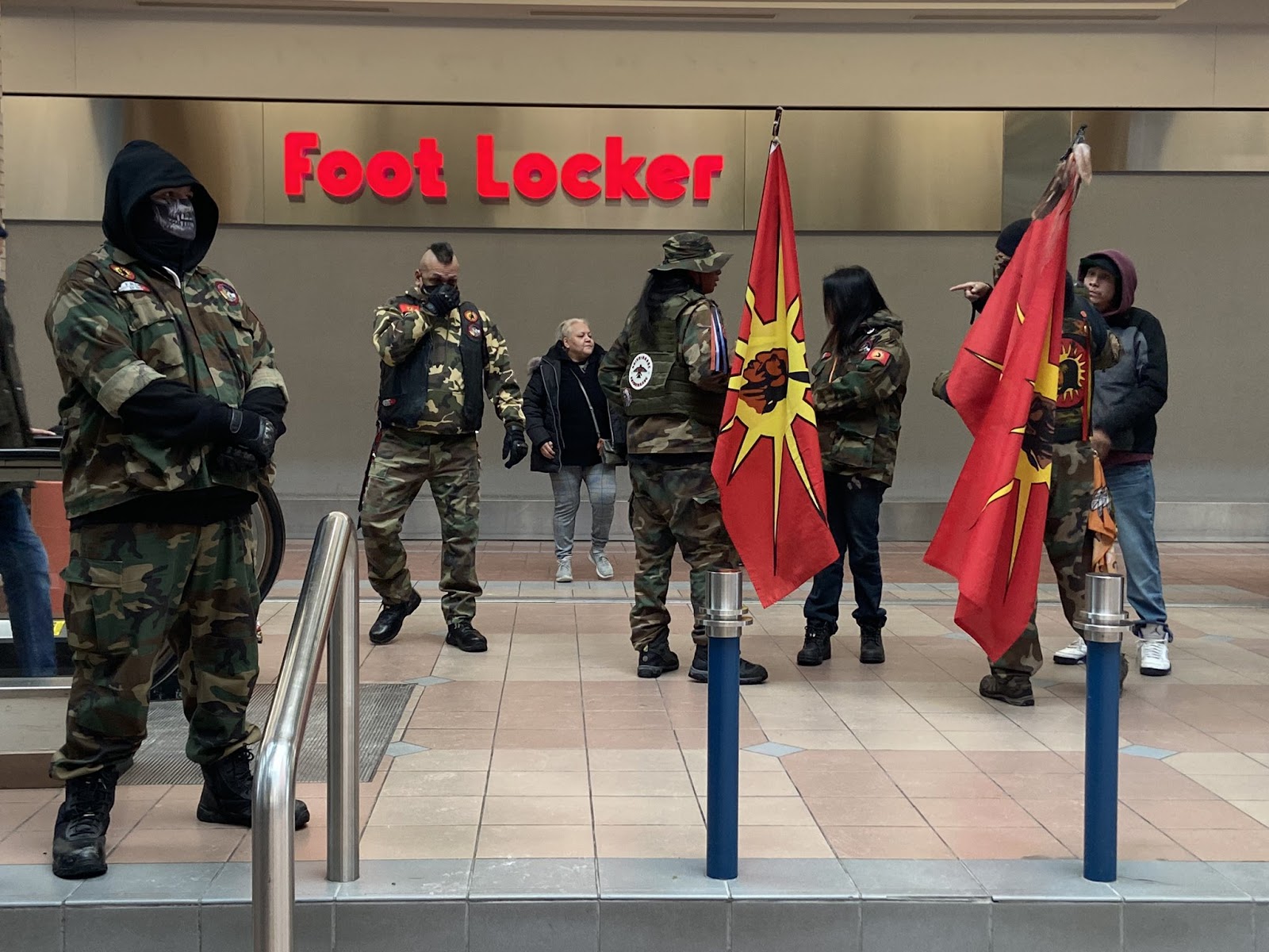 Indigenous activists in camouflage and face coverings gather with flags in Portage Place mall. 