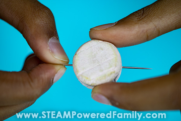 A child's fingers show how to insert the needle into a cut off wine cork while making a homemade compass