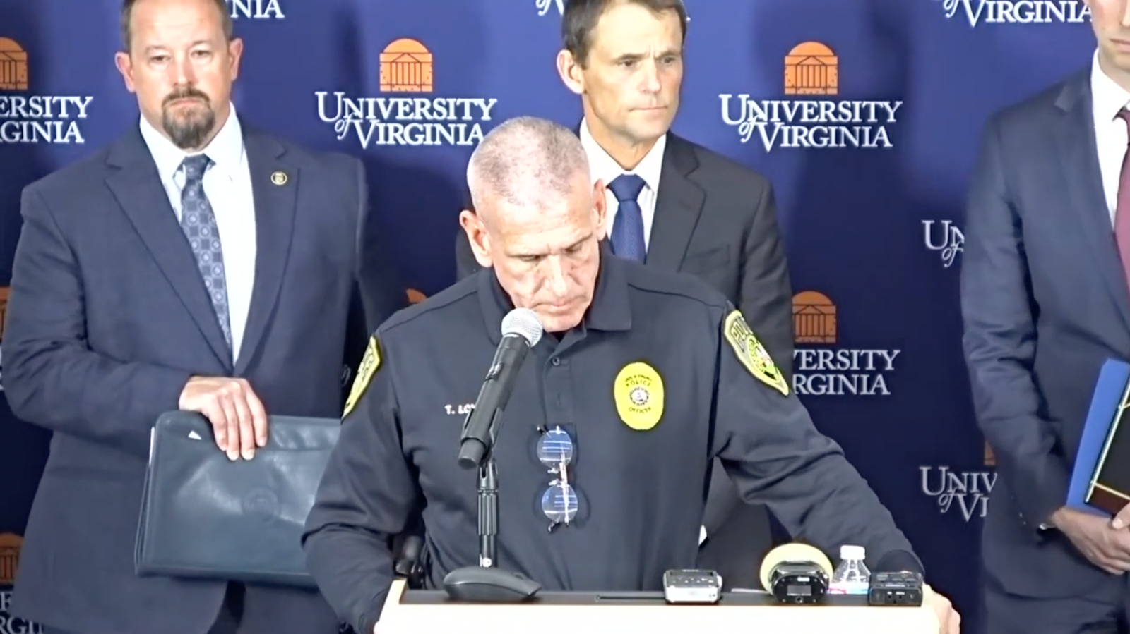 A man in a police uniform stands at a podium with a microphone. Behind him several men stand in suits holding folders.