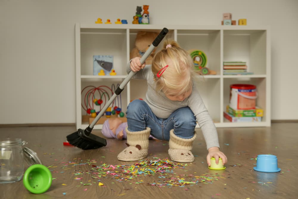 Toddler using broom to clean up spilled confetti in playroom