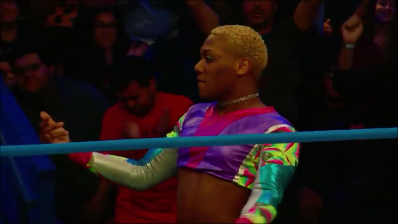 Screen cap from Lucha Underground. Xo Lishus, a black wrestler who presents femme and is wearing a cropped top with neon colors, is facing his opponent who is offscreen. Xo looks cocky and confident. 