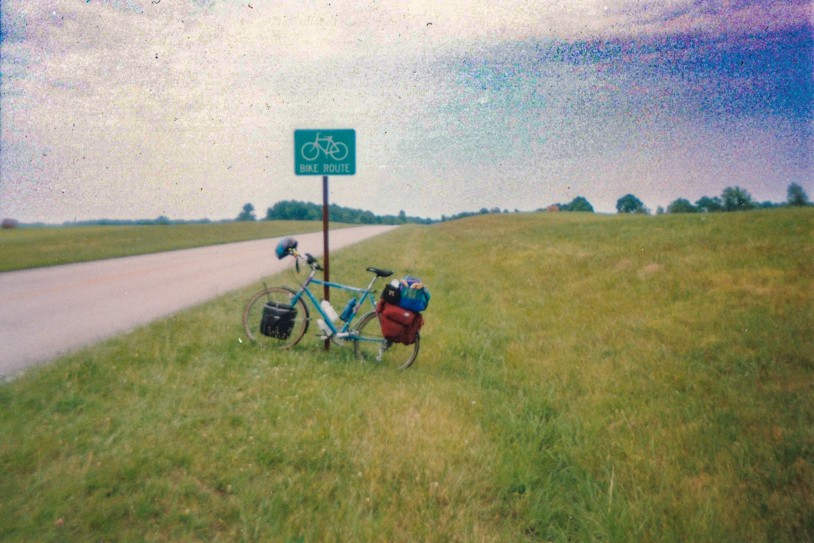 A bicycle packed for camping leans against a sign that notes the road passing through the left side of the photo is a bike route. 