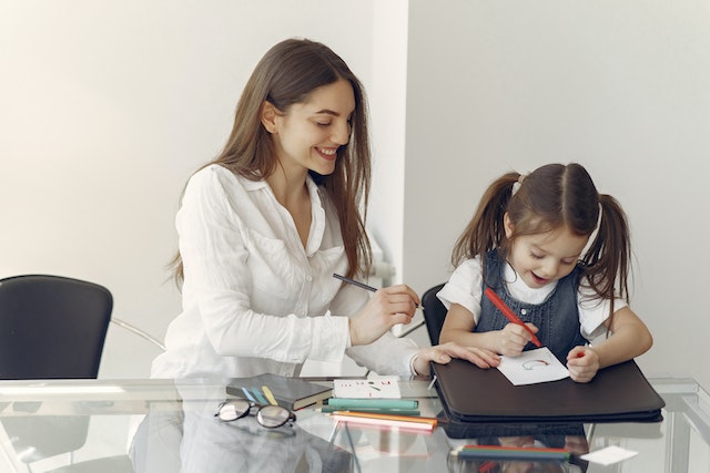 Young girl coloring a picture and a woman sitting beside her at a glass table.