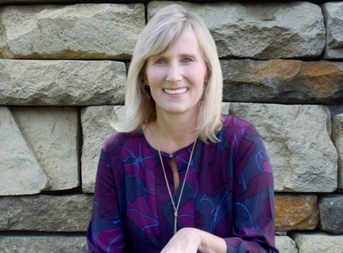 A person smiling in front of a stone wallDescription automatically generated with medium confidence