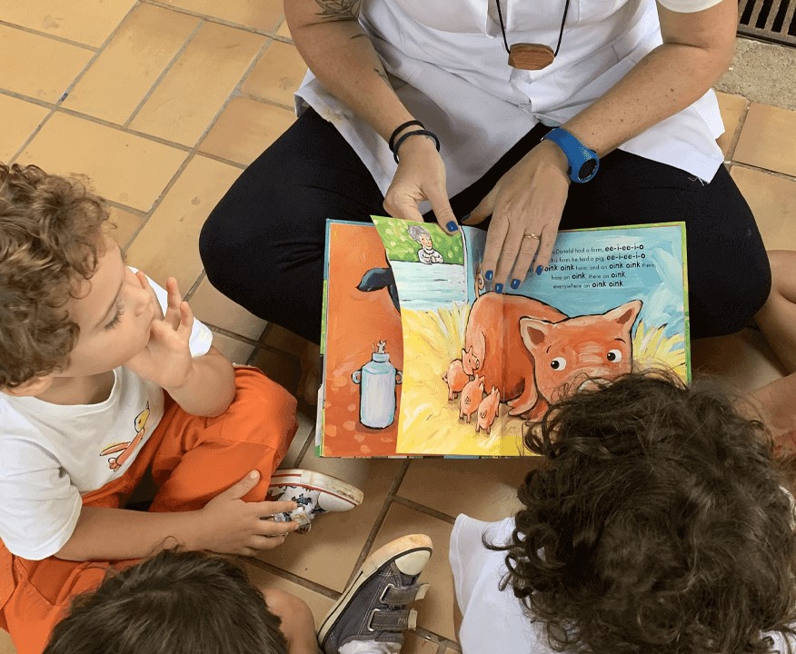 A imagem mostra um adulto mostrando um livro para crianças.