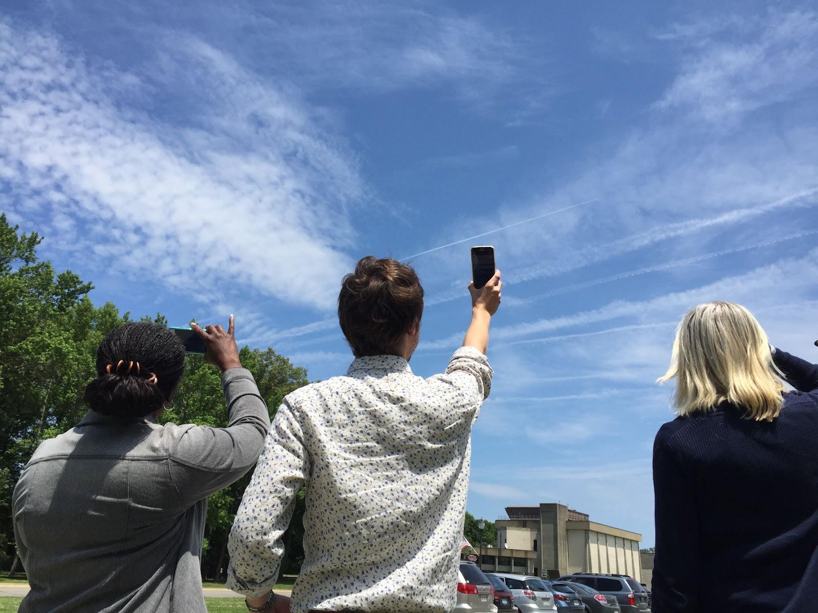 The image has three people, one with braids and dark skin holding a phone, the second has short brown hair and light skin also holding a phone, and the third person has long white hair. All three people are looking at a cloudy sky with contrails and thin cirrus clouds.