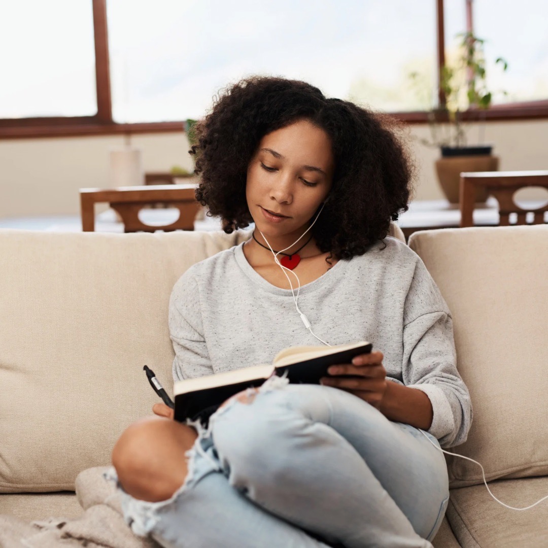 black woman with natural hair, sitting on a couch, using morning journal prompts to write in a journal