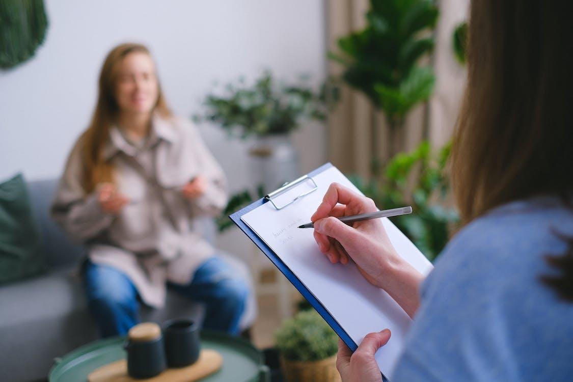 Free Unrecognizable professional female psychologist writing on clipboard while sitting against client on blurred background during psychotherapy session in light office Stock Photo