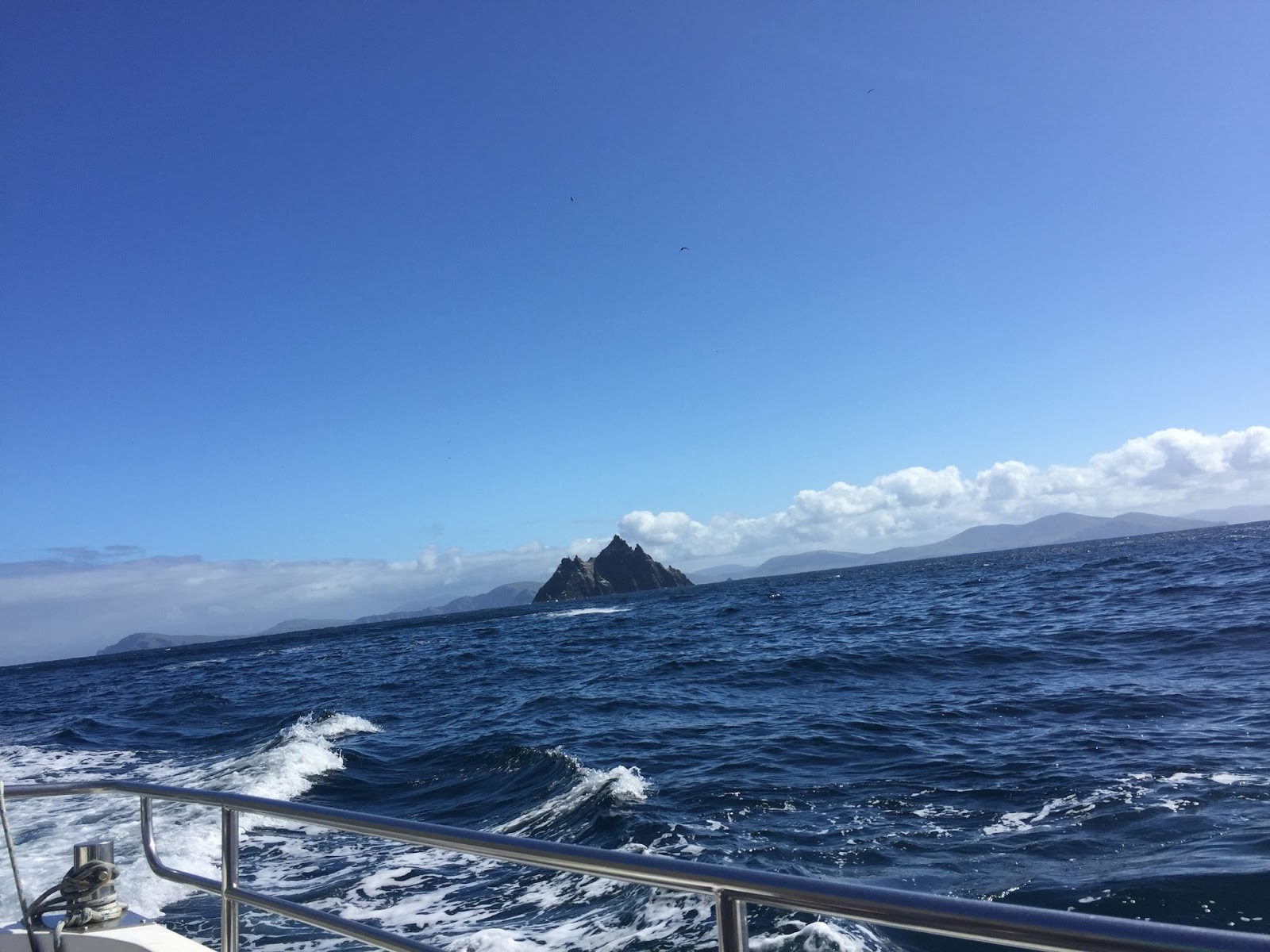 The Skelligs can be seen in the middle distance, jagged rocks rising out of the deep blue sea. The sky is blue with some light fluffy clouds along the horizon. The boat from which the photo is taken appears in the foreground, and the horizon slopes to the left of the photo.