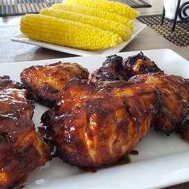 a white ceramic platter holding appealingly sticky-looking grilled chicken coated with barbecue sauce, with corn on the cob in the background