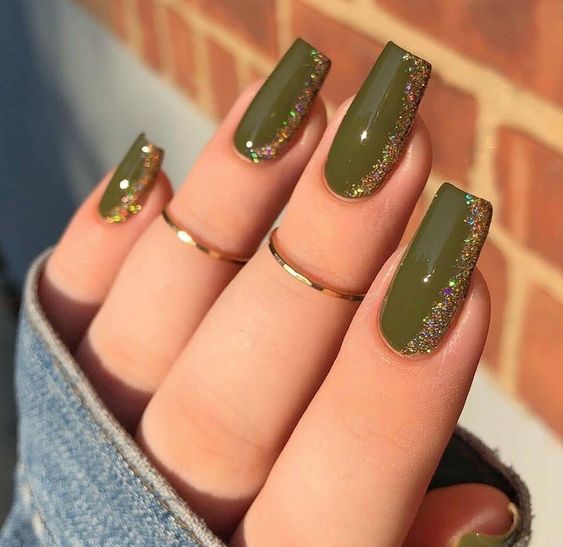 Imagem de uma mão com unhas longas, quadradas e decoradas em verde militar e glitter. A modelo está vestindo anéis dos dedos.
