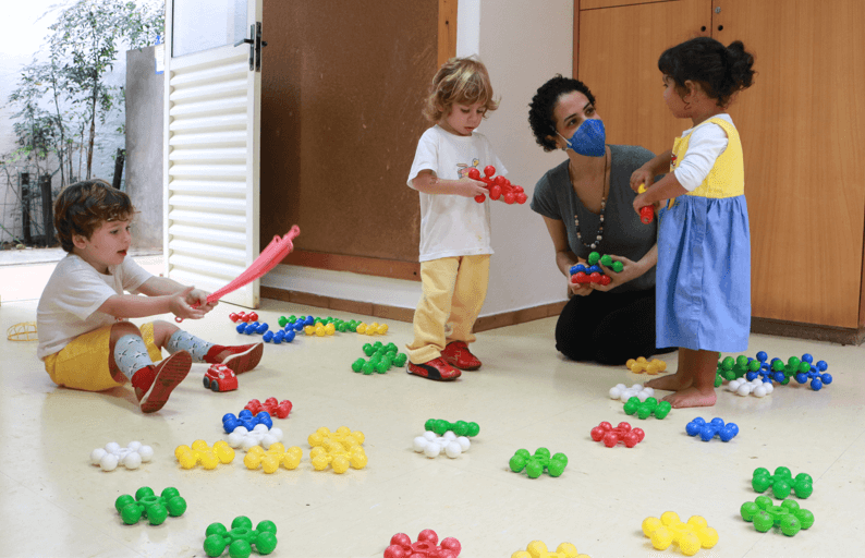 A imagem mostra 1 adulto e 3 crianças em um ambiente com peças coloridas.