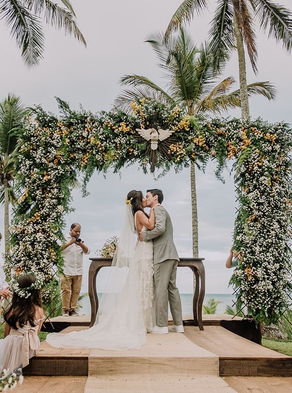 Casamento em Trancoso, uma praia localizada na Bahia. Há um mini altar, montado sob a areia de frente ao mar. Há um arco quadrado de flores brancas com folhas verdes, e o casal está no centro se beijando. A noiva está com um vestido branco rendado, com um véu grande. Ela é branca e possui o cabelo preto. O noivo é branco e possui cabelo preto também, e usa uma roupa social na cor cinza. 