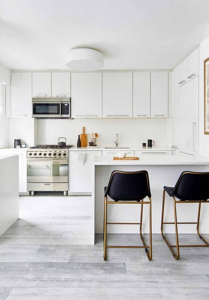 Cozinha com armários brancos, eletrodomésticos de inox, piso de madeira branca, bancada branca com cadeiras pretas e detalhes dourados.