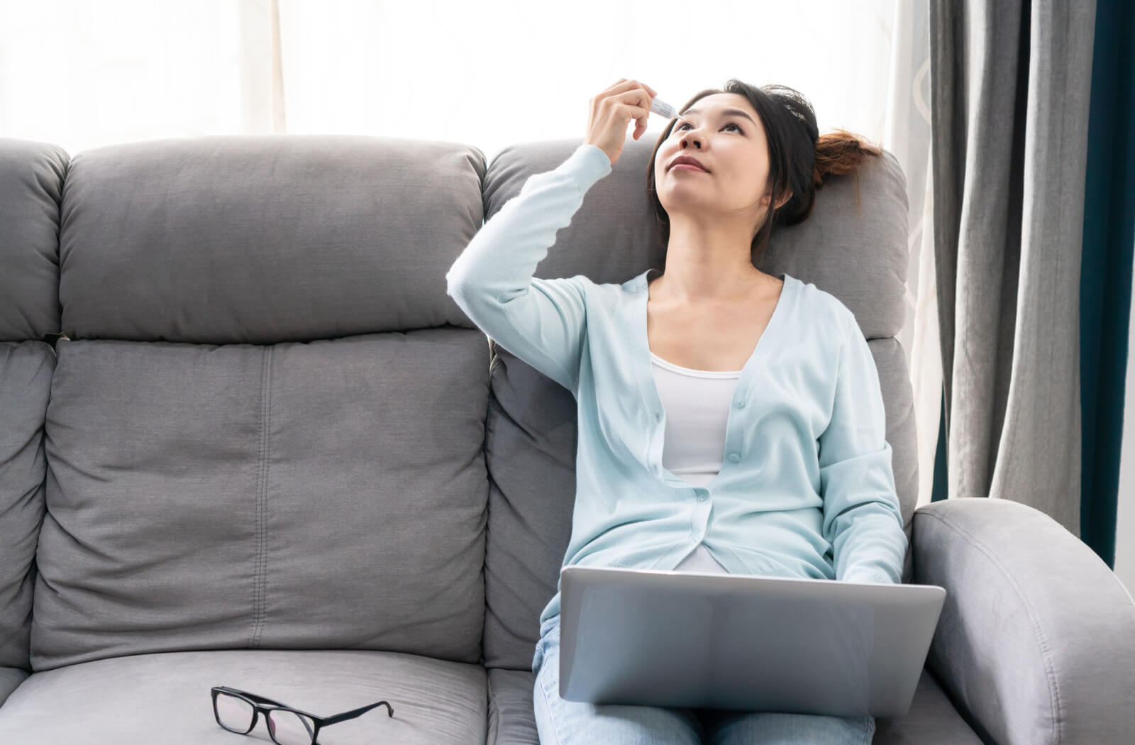 A woman in a blue cardigan applying eye drops on her right eye while working on her laptop.