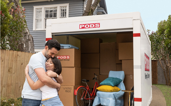 A happy couple is smiling and embracing in front of their loaded PODS portable moving container. It’s filled with neatly stacked moving boxes, a chair wrapped in a moving blanket, throw pillows, and a bicycle.