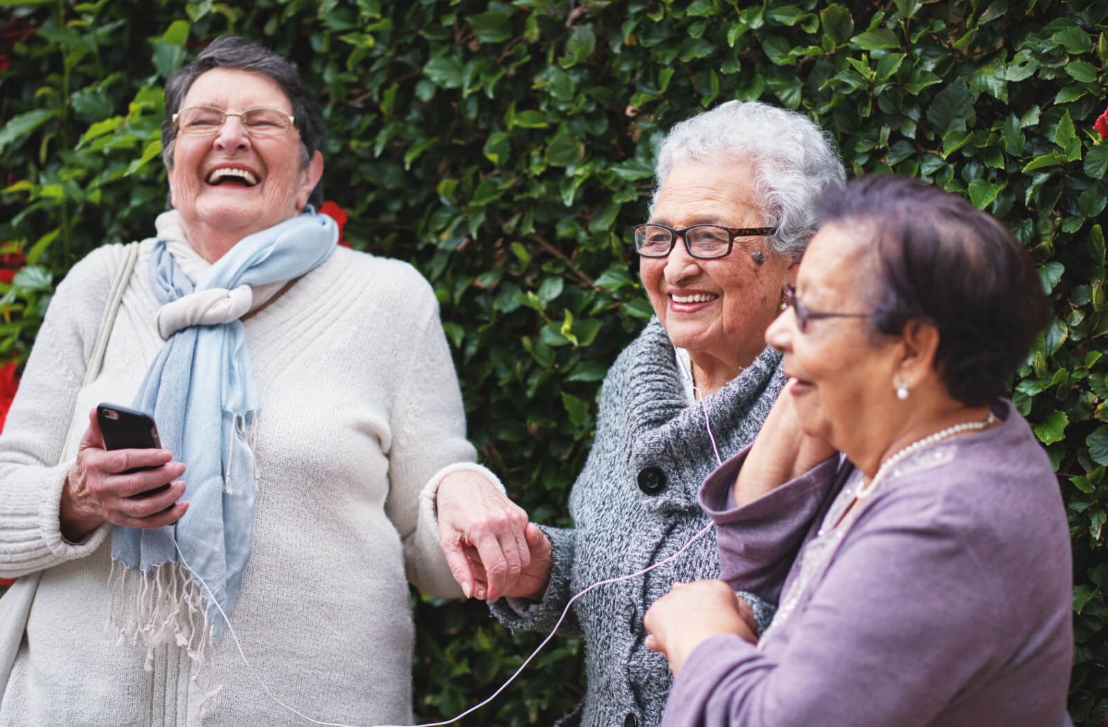 A group of senior women smiling and listening to music on a smartphone while wearing earphones.