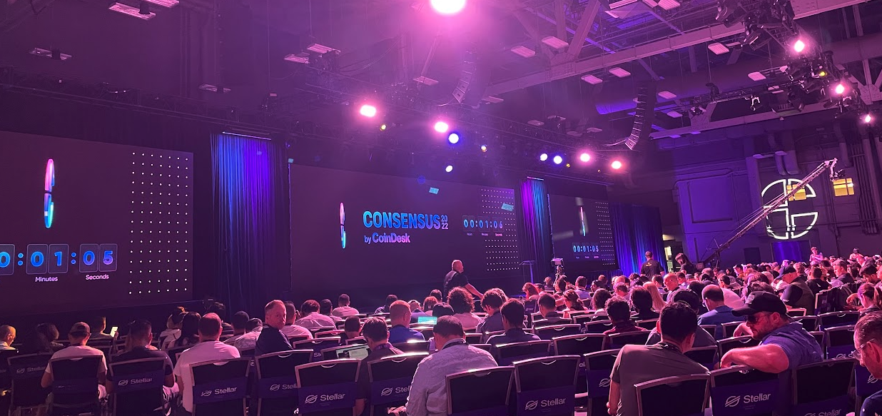 Foto do palco principal do evento Consensus 2022. Na imagem, aparece a contagem regressiva em um telão, luzes no tom roxo e muitas pessoas sentadas aguardando o início do evento.