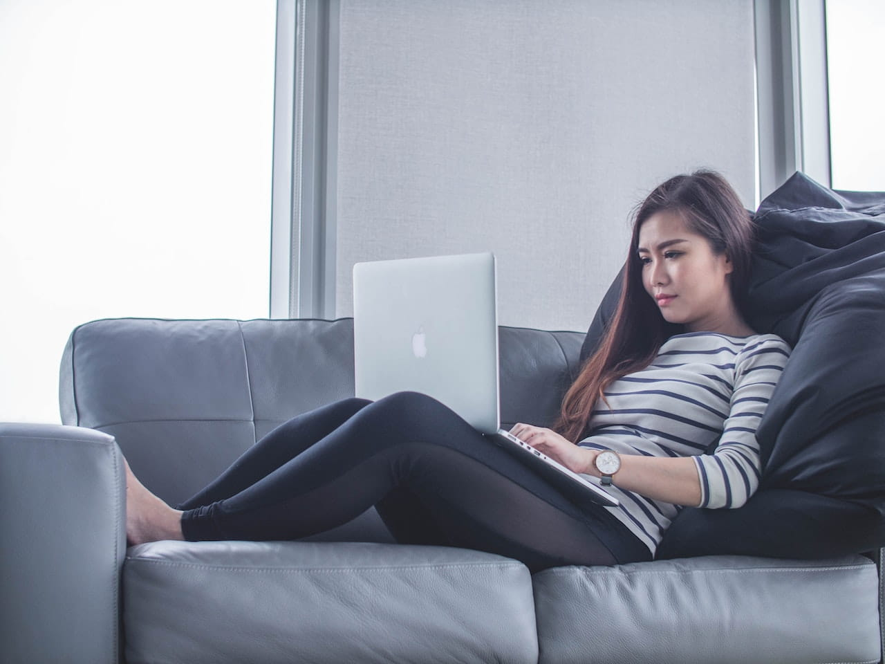 A young woman lays on a couch while working on her laptop.