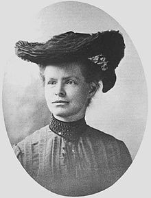 A old black and white photo shows a woman looking into the distance wearing old fashioned clothes and a hat.