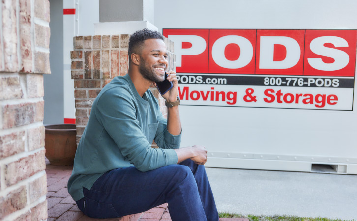 A man sits on his front step while he talks on his phone near a PODS moving and storage container in his driveway