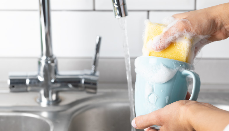 Someone using a soapy sponge to wash a light-blue mug in a kitchen sink