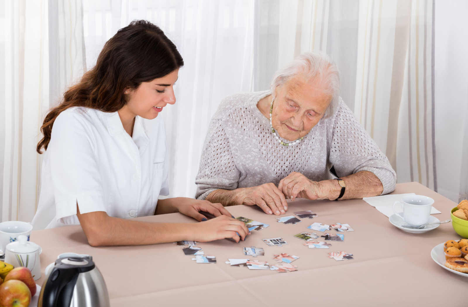 A young female nursing assistant in her white uniform is sitting beside an old lady while they are playing puzzles on the table.