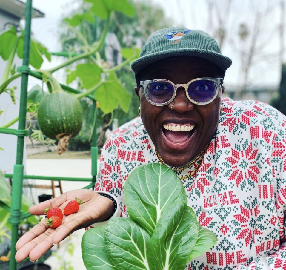 Shantira Jackson smiles widely with three small strawberries in her hand. She wears a green corduroy baseball cap, glasses, and a colorful top. She’s a rising star in the comedy game with writing credits on hit shows like Big Mouth and the Amber Ruffin Show.