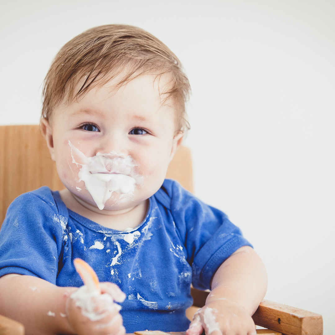 Baby spoon feeding himself a bowl of yoghurt and making a mess