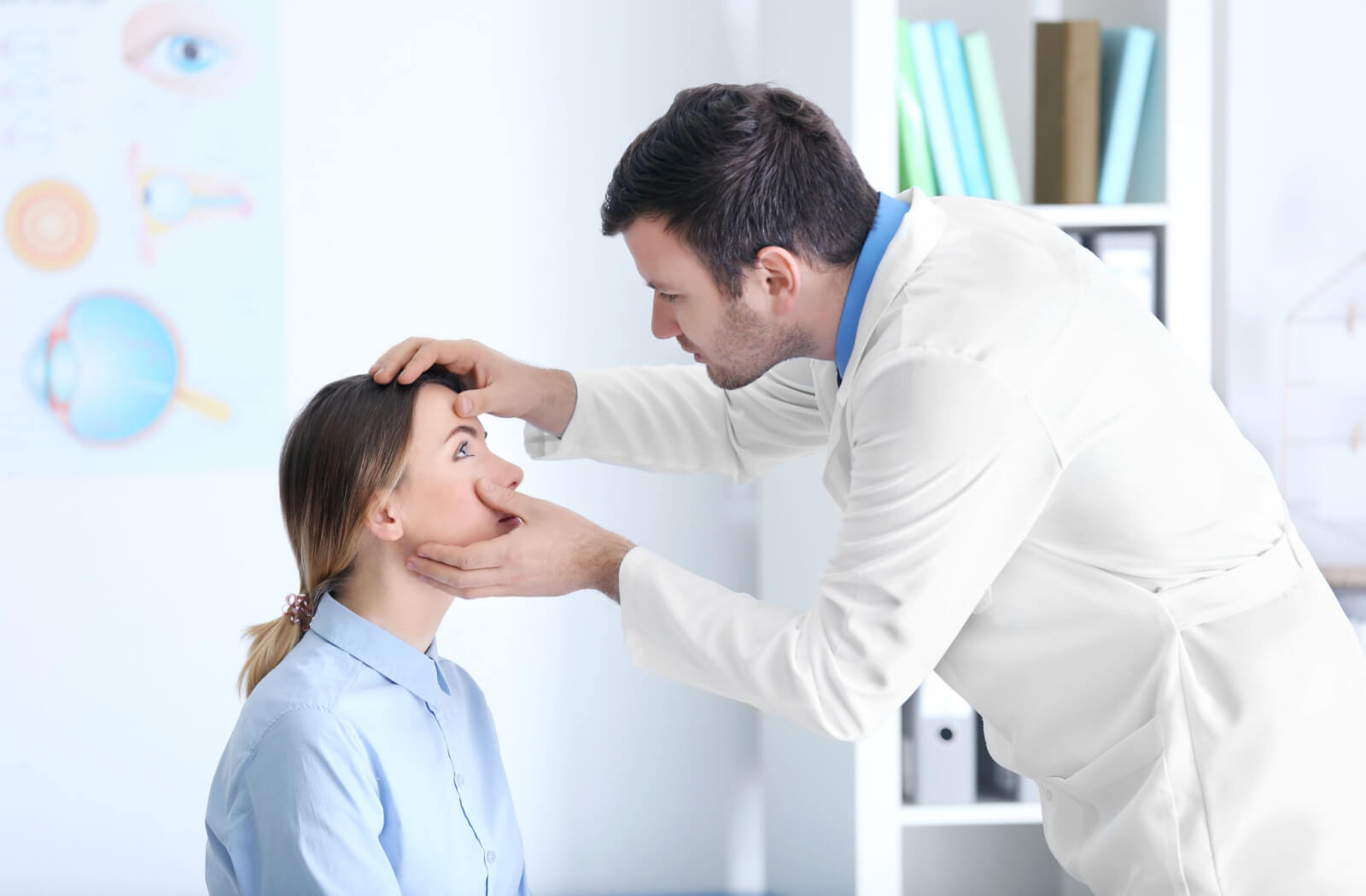 A male eye doctor is examining the eye of a female patient in a light blue collared long-sleeve for her regular eye check-up. The doctor is looking closely at the eye of the patient.