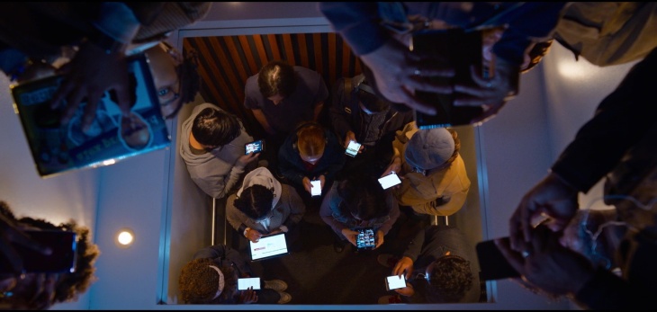 Image of several people in an elevator all looking at their phones - except Neo.