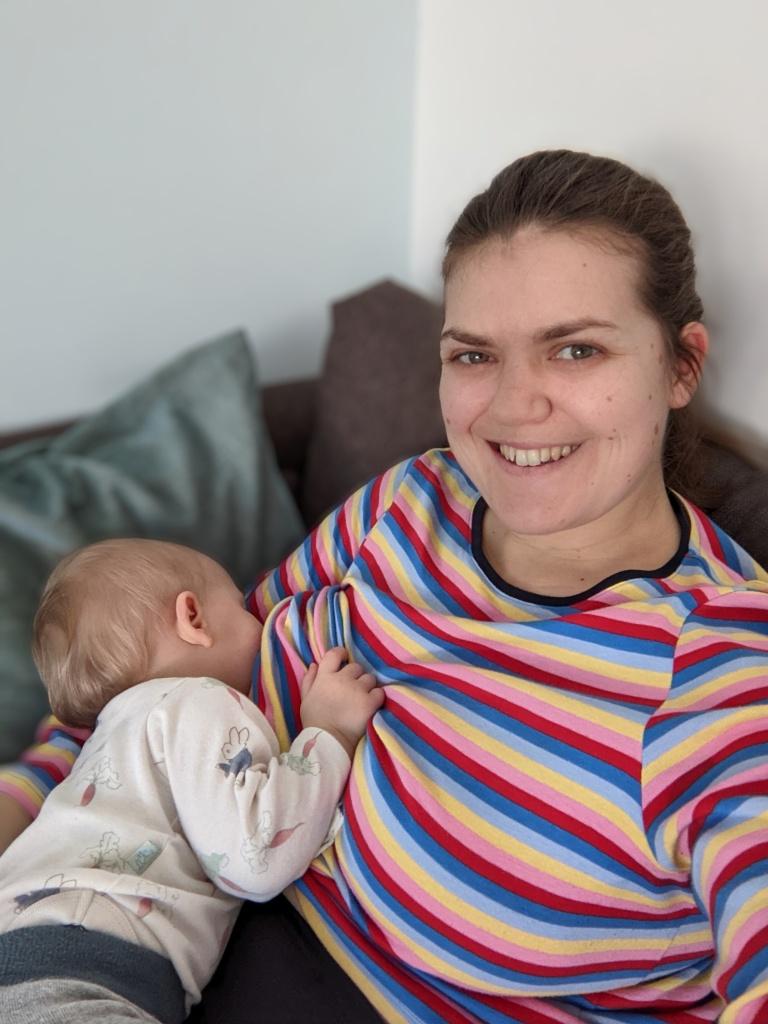 Fiona is wearing a bright, stripy top, sitting on a grey sofa feeding her daughter who is napping while laying on her lap. Her daughter is wearing a Peter Rabbit top and grey leggings.