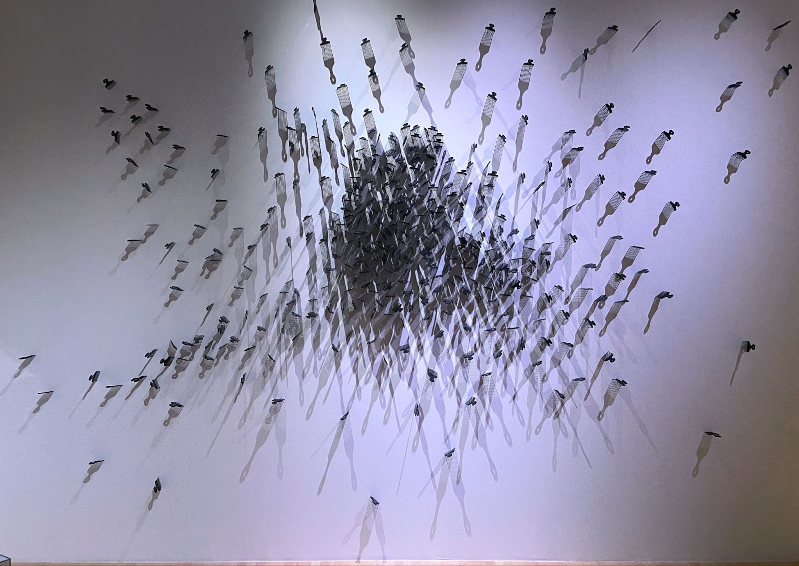 Image: A wall installation of black hair picks embedded in a white gallery wall. There is a larger cluster of combs in the center of the image, with other combs fanning out in a vague star-shaped pattern. Photo by Jessica Hammie. 
