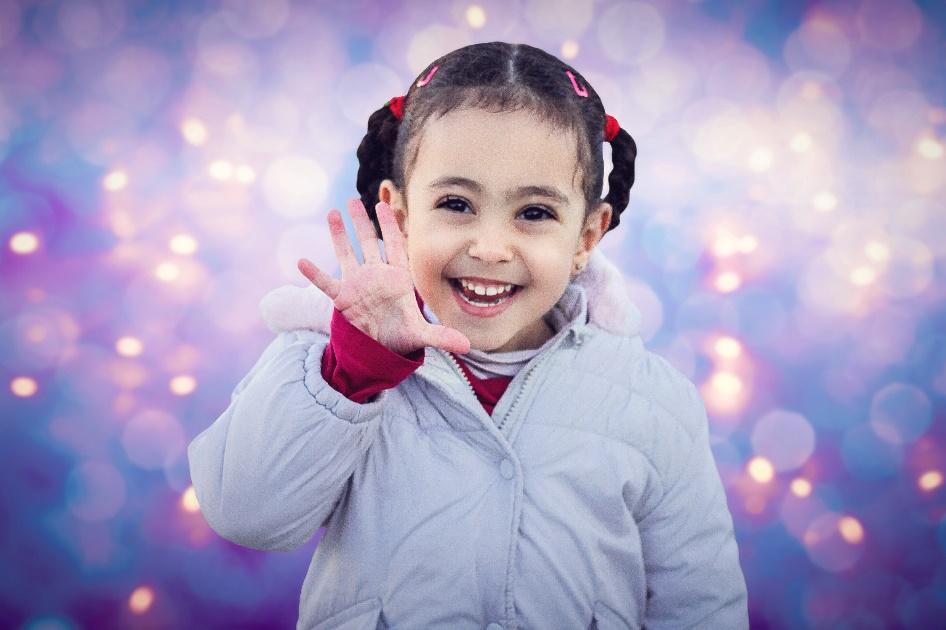 Young girl waves to the camera, smiling against a background of pixilated purple, pink and yellow light. 