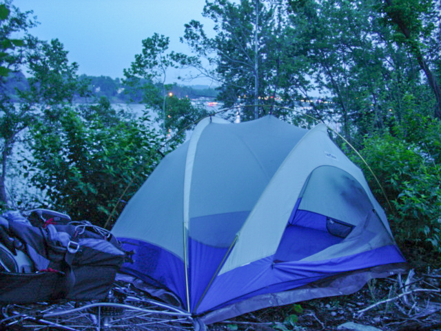 At night a tent is visible in the cover of trees. A river and lights are in the background. 