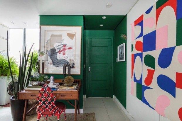 Ambiente com hall de entrada e porta verde escuro, demais paredes brancas com desenhos coloridos, piso branco e escrivaninha de madeira com cadeira vermelha.