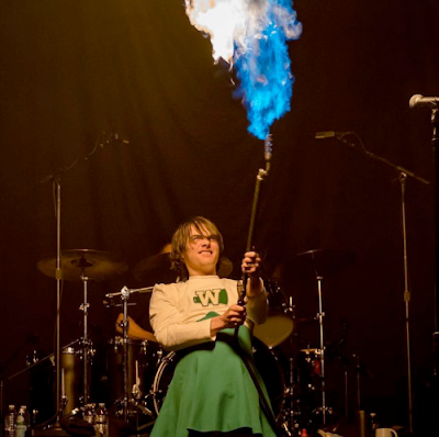 Gerard again, in his 40s again. He is standing onstage in front of a drum kit. He is wearing a green-and-white, vintage, women's-style cheerleading uniform with a "W" emblazoned on the front. His brown hair falls loosely around his face. He is wielding a flamethrower, shooting fire high into the air above him. He is grinning like a little kid.
