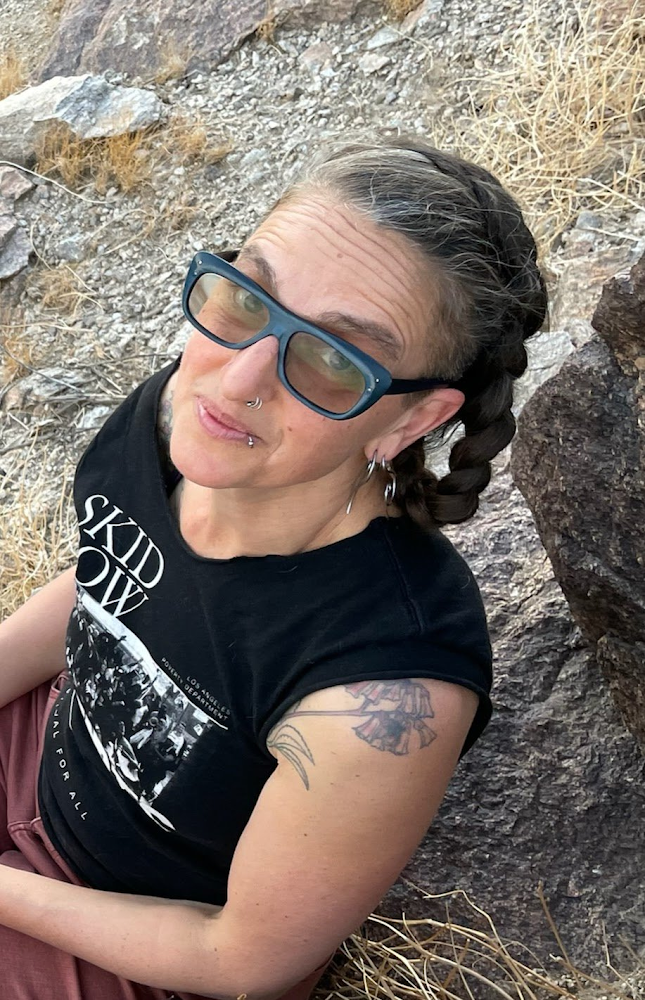 Image Description: A white Latinx Jewish queer person with long brown hair in a braid, streaked with silver. They are wearing light-colored shades with blue frames, and have multiple facial piercings. They are sitting on rocky earth, looking up toward the camera with raised eyebrows. They are wearing a sleeveless Festival for all Skid Row Artists t-shirt and dusty rose-colored pants. A tattoo of a flowering succulent is visible peeking out of their shirt on their left shoulder.