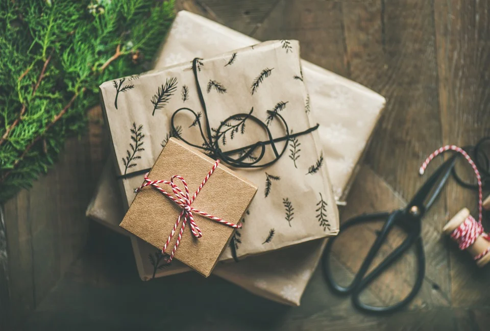 3 paquets cadeaux gris et marrons, entourés de rubans cadeaux posés sur un parquet de bois marron avec un ciseau et une branche de sapin en arrière plan