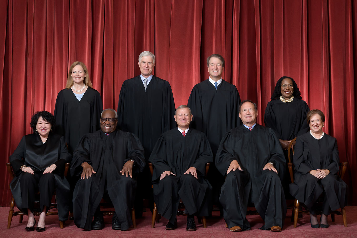 Foto dos nove juízes da Corte americana usando uma toga preta em um fundo acortinado vermelho; quatro estão em pé e cinco estão sentados