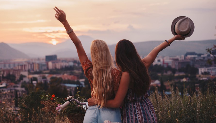 photo of two millennial girls throwing hats off over a hilltop view 
