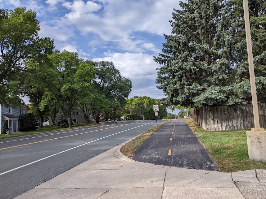 Asphalt pathway along a roadway lined with houses with evergreen and deciduous trees along the front yards. The yard on the side of the road with the trail has a wooden lathe fence along the yard.