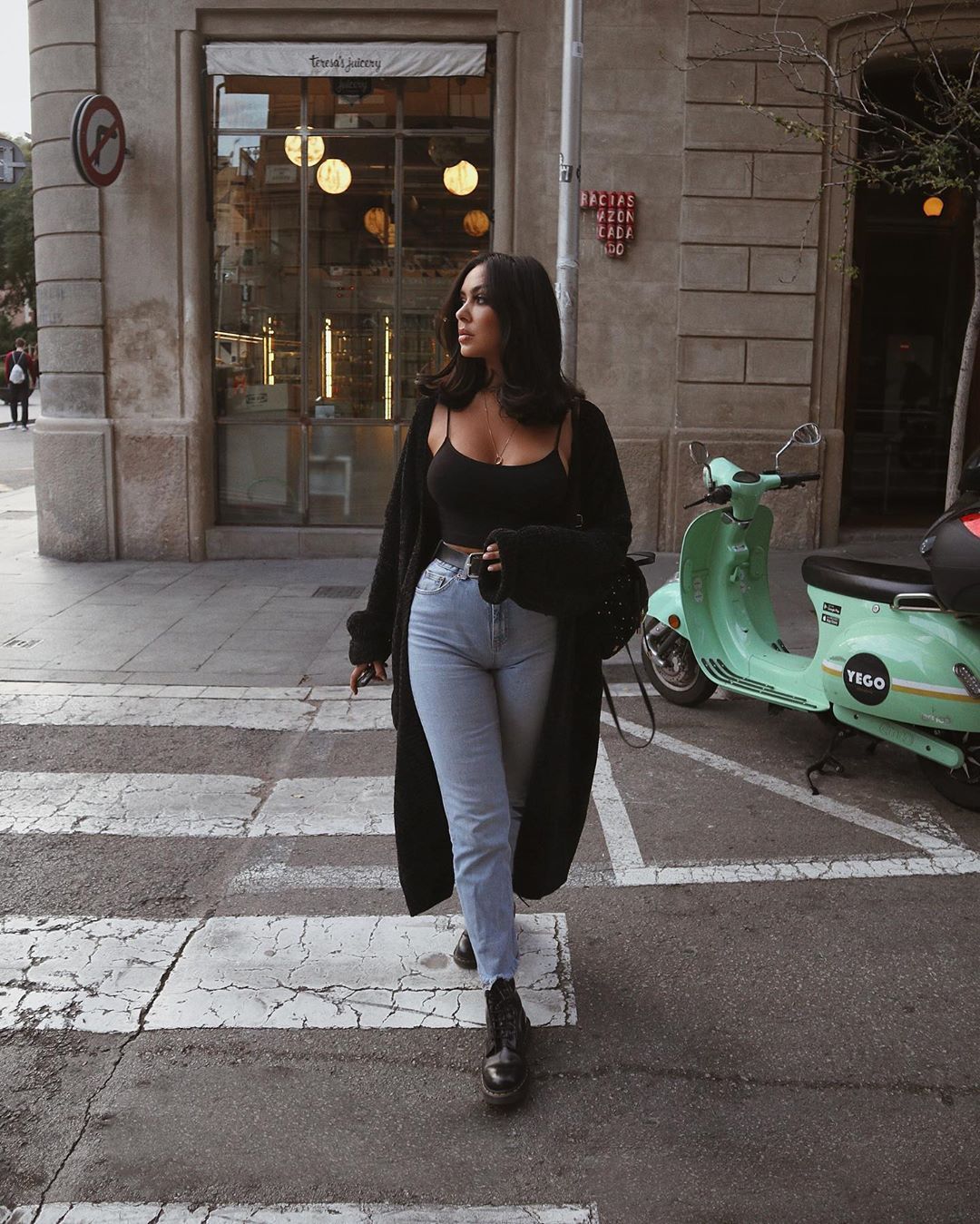 Mulher branca, de cabelos e olhos castanhos atravessando a rua. Ela usa um coturno baixo, uma calça jeans clara, blusa simples de alcinha preta e um casaco preto. 