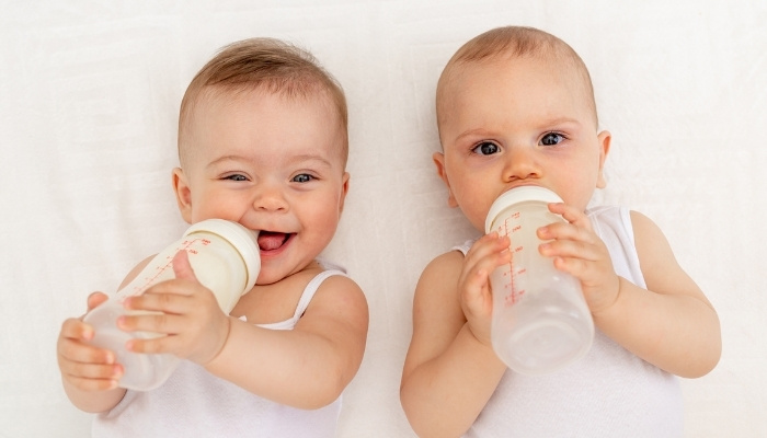 Baby twin sisters holding their own bottles with one smiling and the other looking serious.